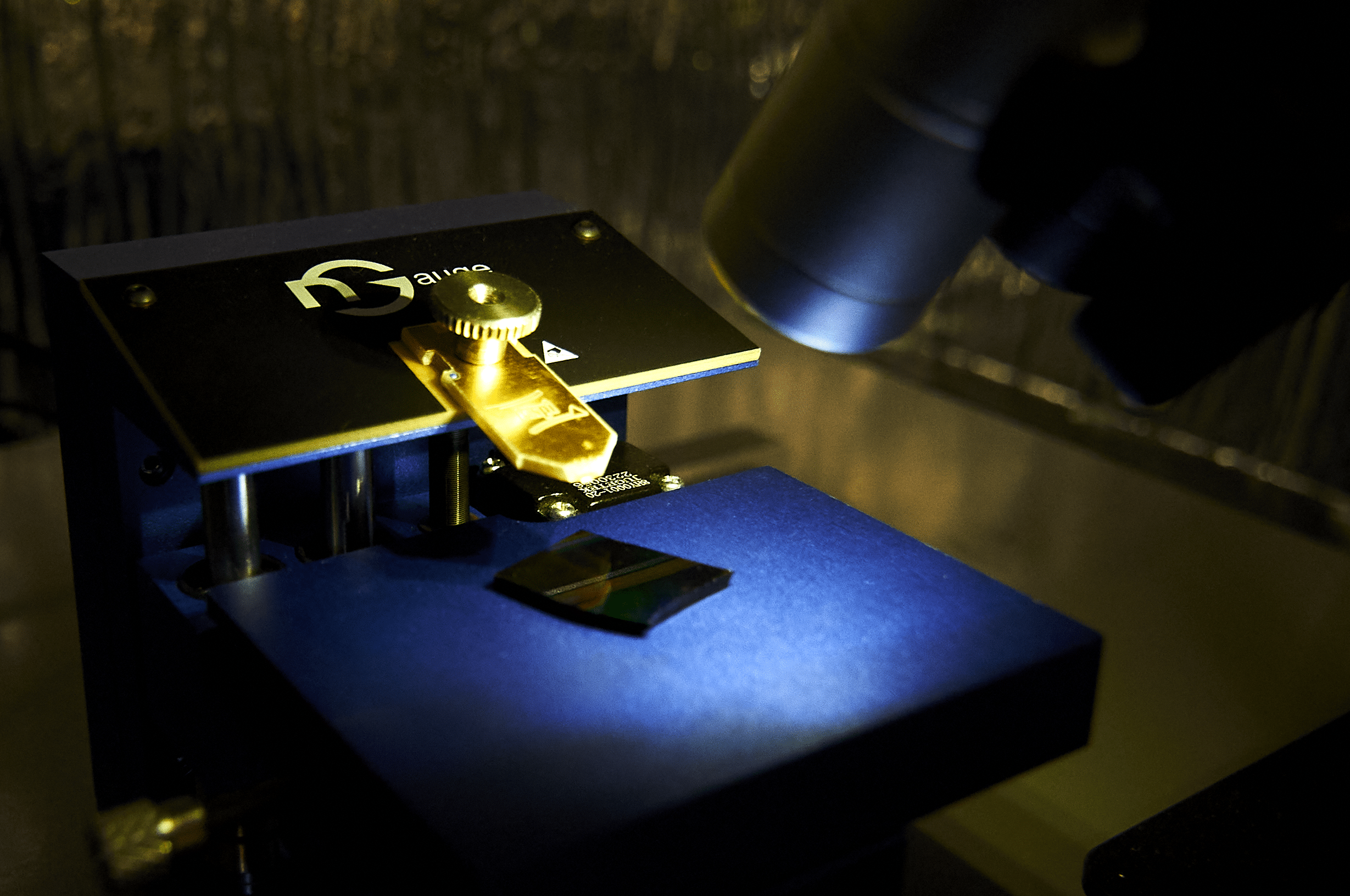 Close-up of a nGauge AFM microscope examining a small rectangular sample on a metallic stage, illuminated under a focused light.