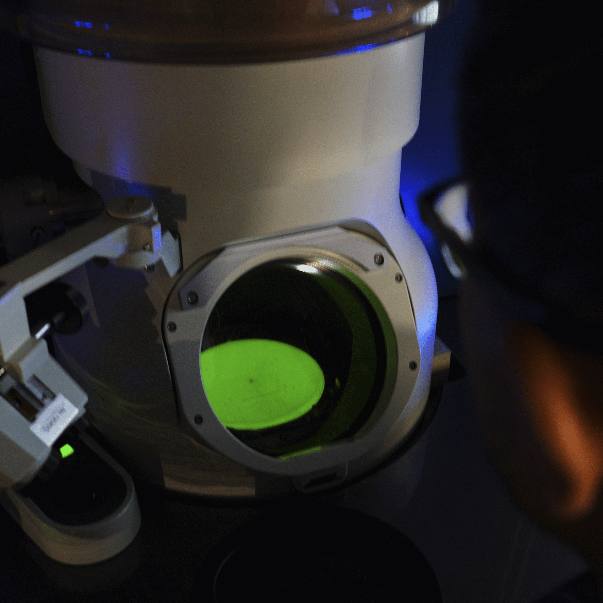 Person observing a sample inside a laboratory electron microscope with a green-lit screen.