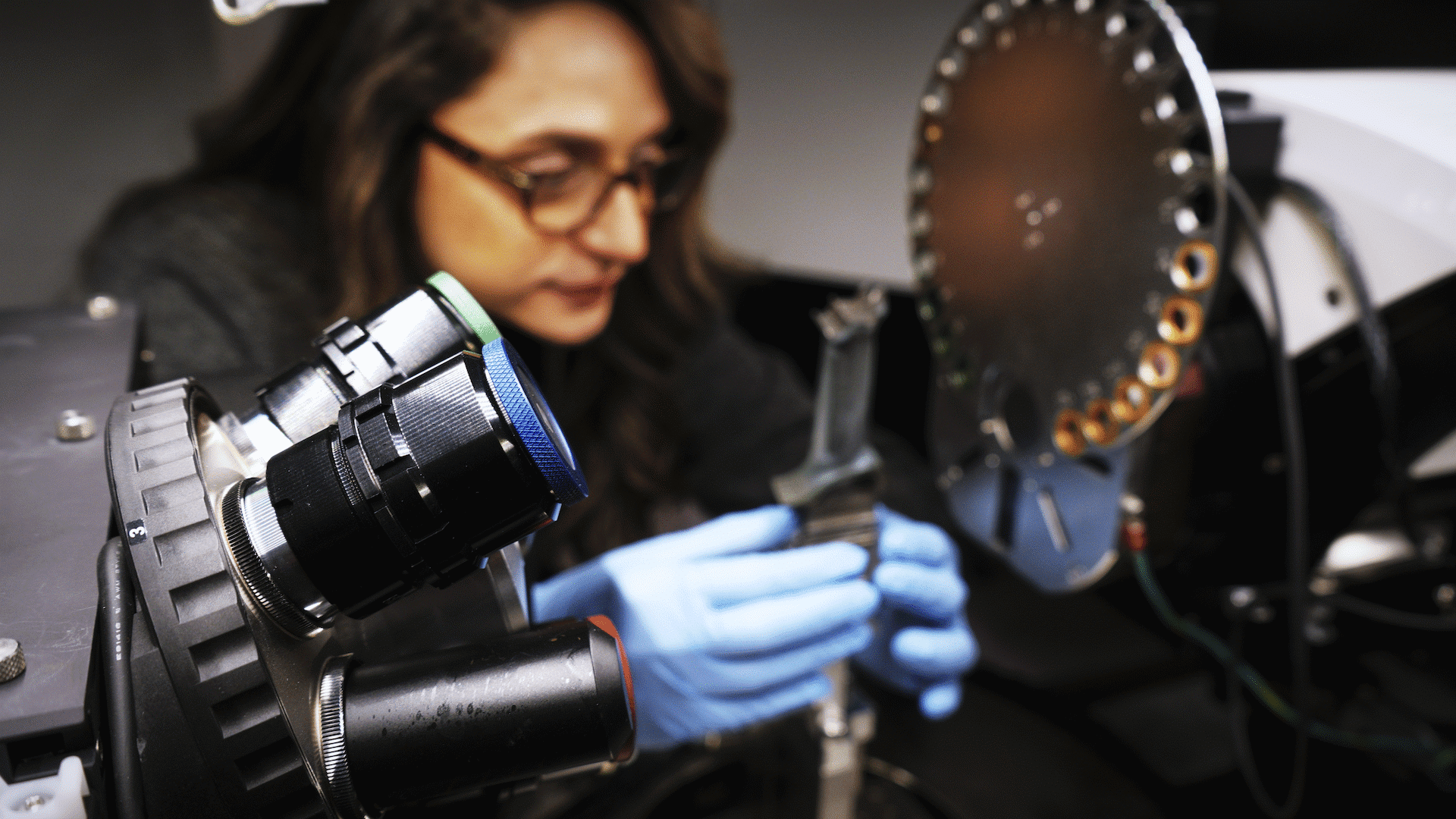A person wearing glasses and blue gloves examines equipment in a laboratory setting.