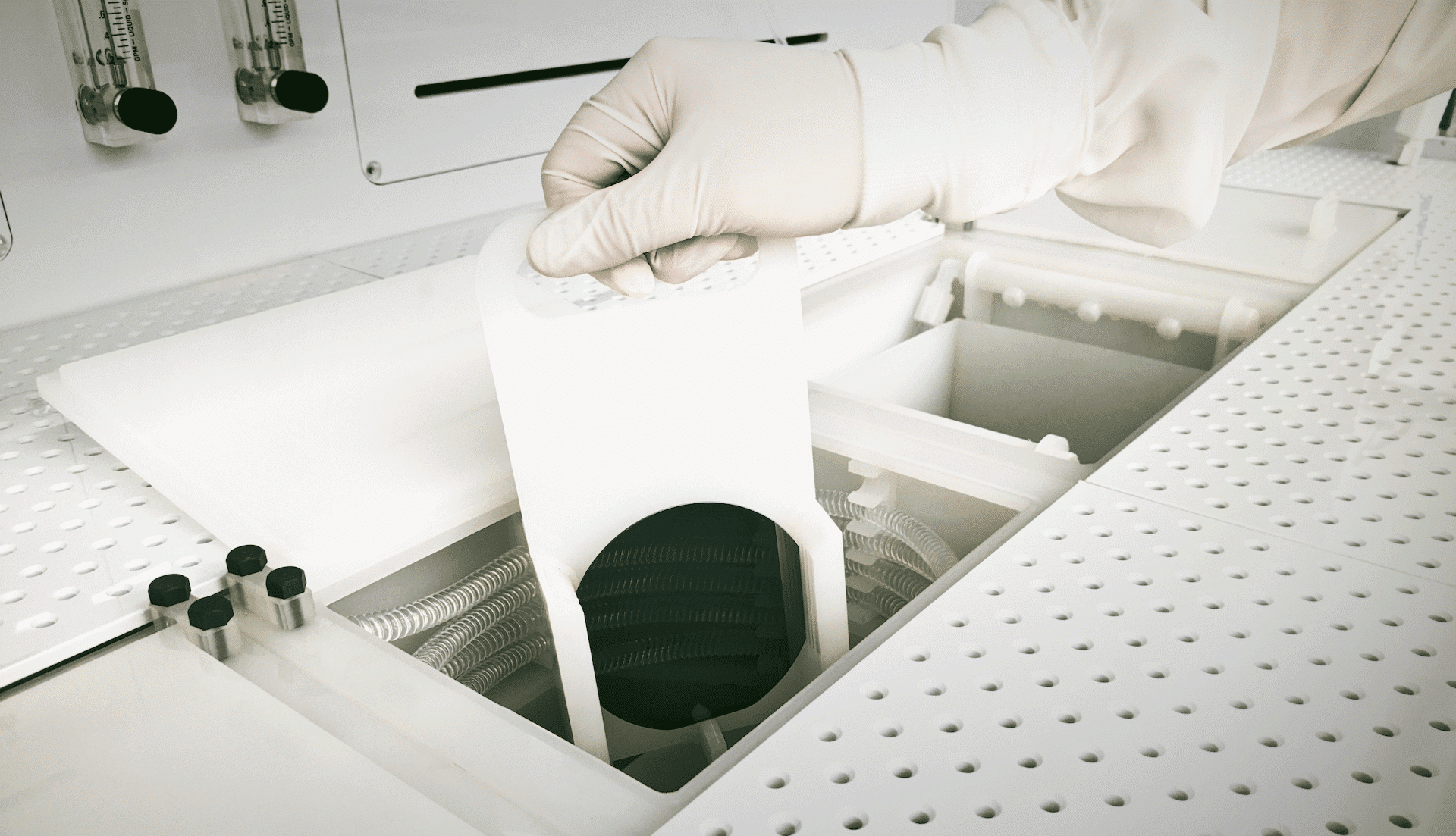 A gloved hand lifts a container holding circular silicon wafers from a white, perforated surface in a clean room setting.