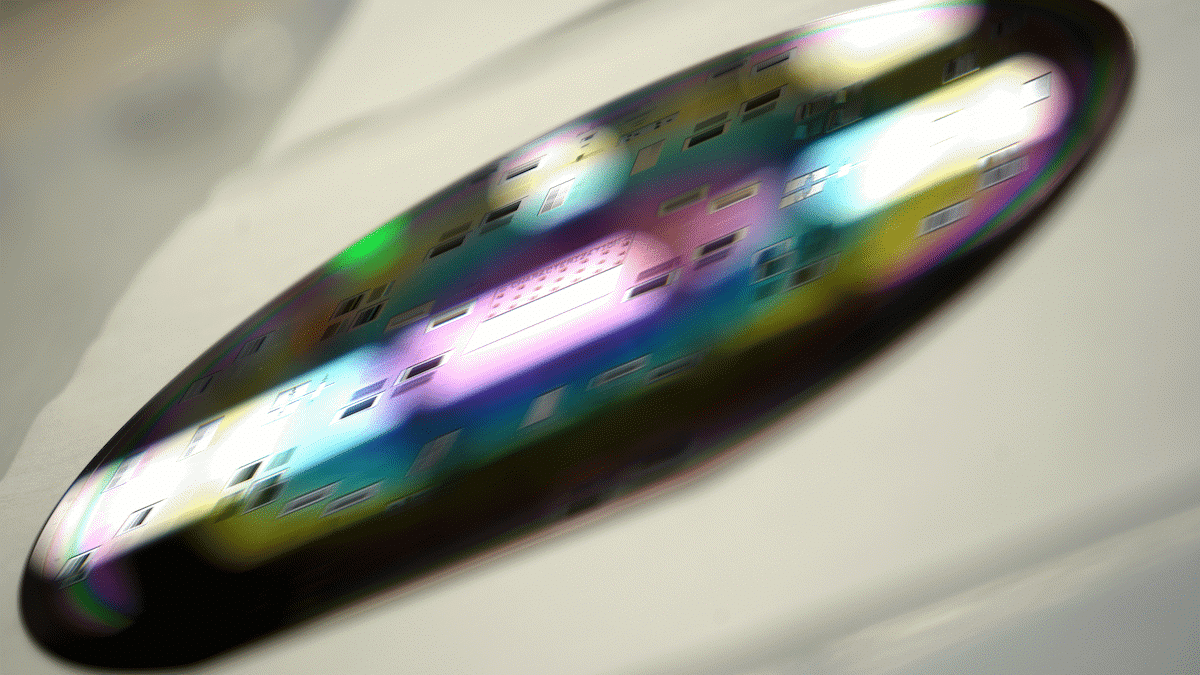 Close-up of a silicon wafer displaying colorful, reflective patterns under light, on a blurred white background.