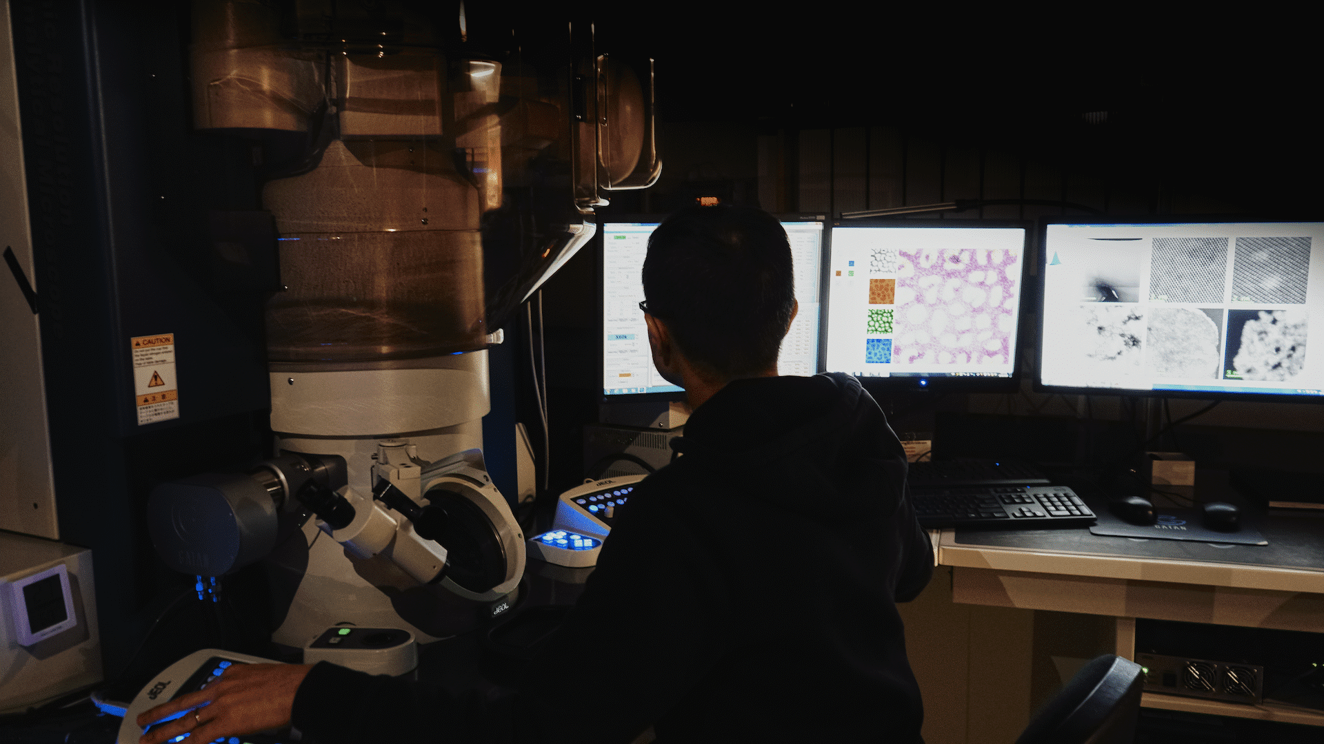 A person operates a large microscope, focused on several computer monitors displaying microscopic images.