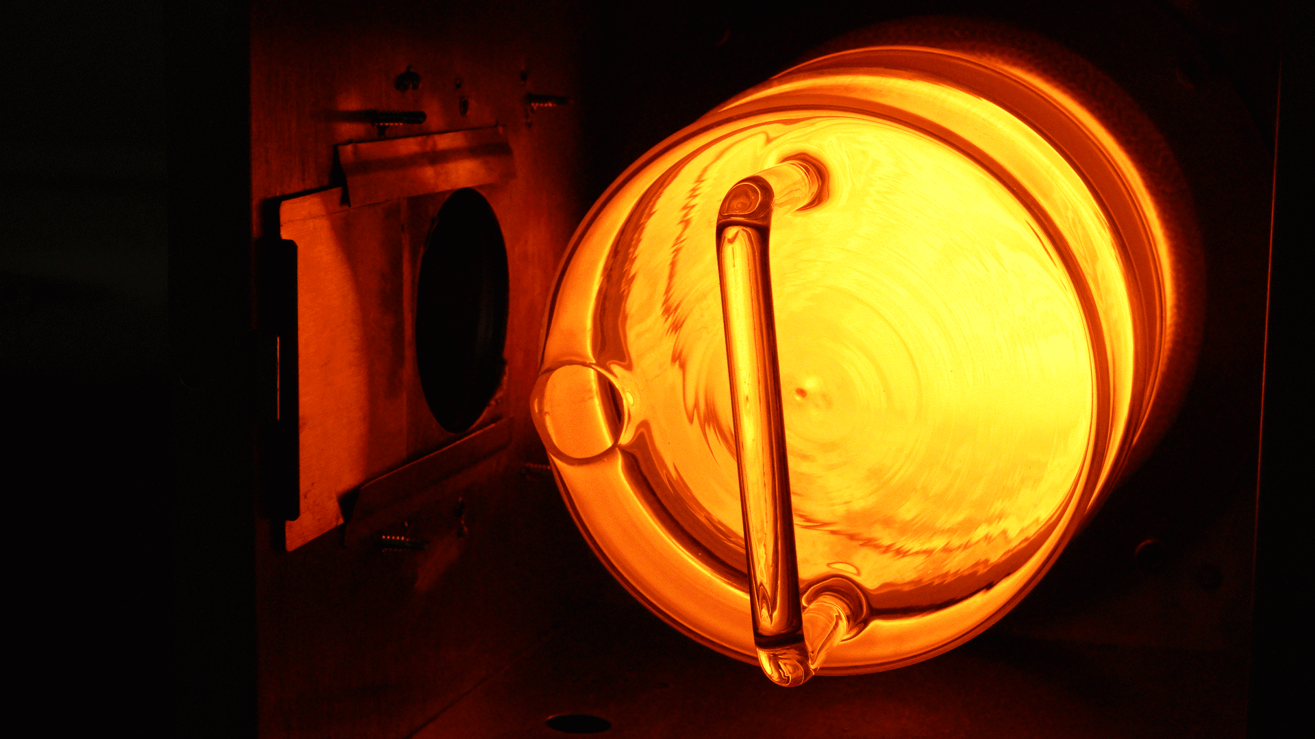 Close-up of a stage spotlight with a warm orange glow. A metal handle is attached to the circular, illuminated front, surrounded by a dark, partially visible housing.