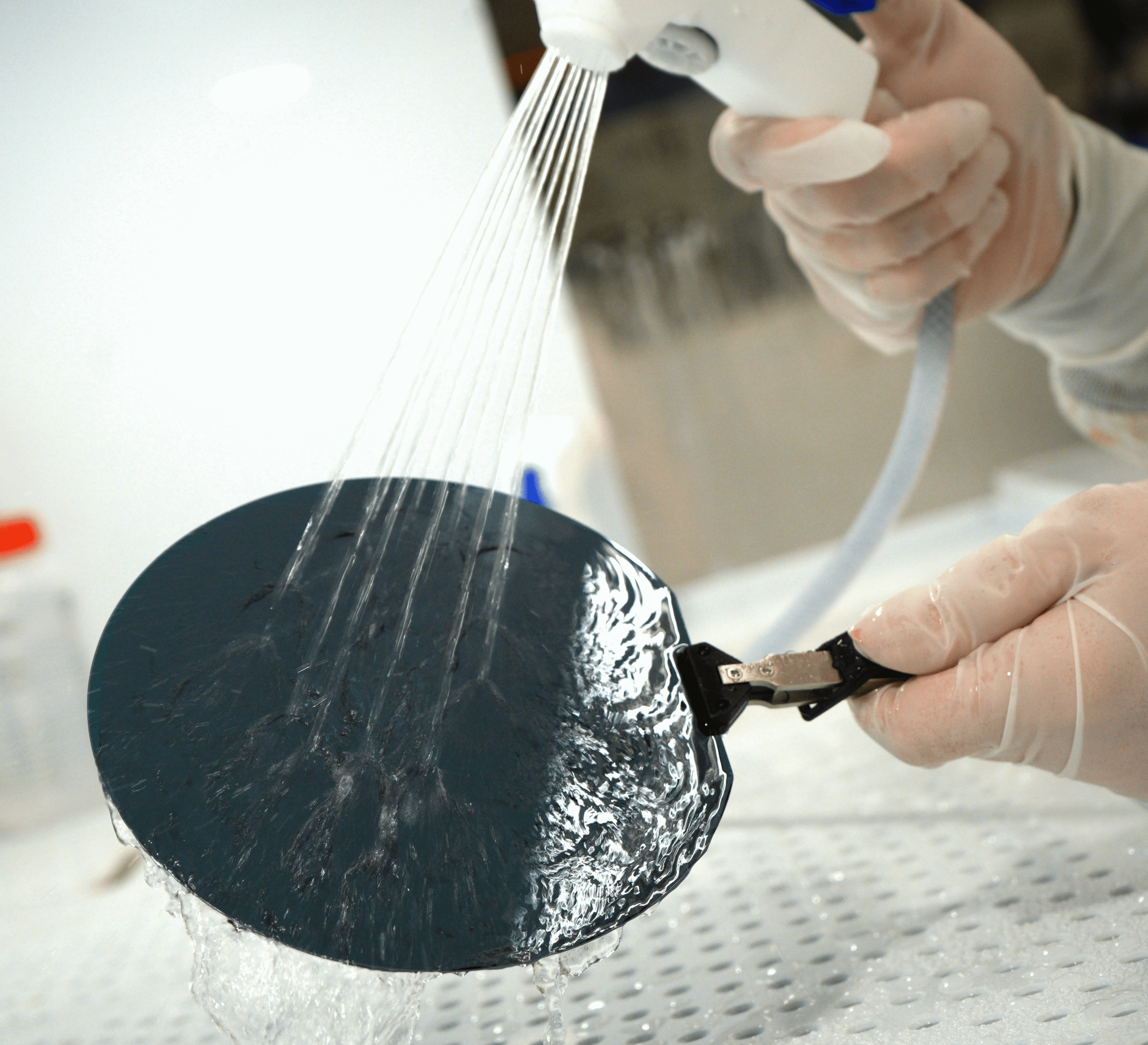 A person wearing gloves rinses a circular silicon wafer with a spray bottle, over a white perforated surface.